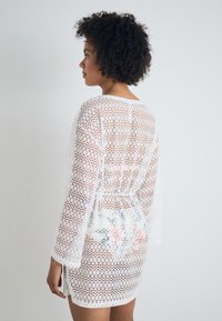 Vestido blanco de ganchillo con mangas largas, que presenta una textura de punto abierto y un acento floral en la parte trasera. Ajustado al cuerpo con una cintura ceñida.