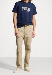 Námořnické bavlněné tričko s nápisem "POLO" a "RALPH LAUREN", v kombinaci s bledě hnědými plátěnými kalhotami se splatterem barvy a bílými teniskami.