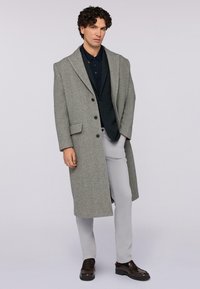 Cappotto lungo in misto lana grigio con ampi rever, tre bottoni e una tasca laterale, indossato sopra una camicia nera e pantaloni grigio chiaro.