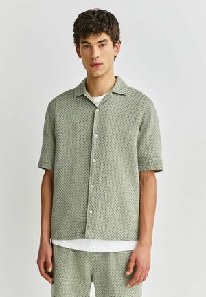 Junger Mann in passendem hellgrün gemustertem kurzärmligem Hemd und Shorts mit weißem Unterhemd, stehend vor einem einfarbigen Hintergrund.