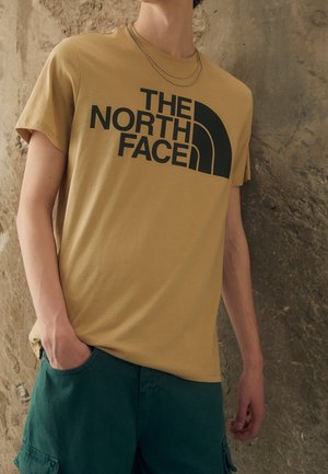 Camiseta de color mostaza con un logo gráfico negro de "The North Face". Mangas cortas, cuello redondo y un ajuste relajado.