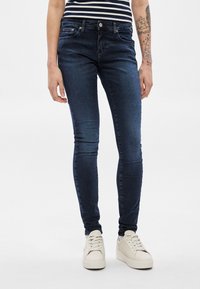 Tmavomodré úzke rifle z denimu. Majú päť vreciek, zips a tónované prešívanie. Skombinované s bielymi teniskami.