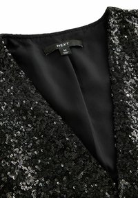 Gros plan sur une veste noire à sequins avec une étiquette de marque « NEXT » et une étiquette de taille moyenne à l'intérieur du col.