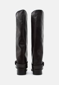 Ζευγάρι ψηλών σκούρων καφέ δερμάτινων μπότες με χαμηλά τακούνια τύπου block και λουριά στον αστράγαλο, εμφανιζόμενες από πίσω σε λευκό φόντο.