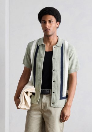 Młody mężczyzna ubrany w jasnzieloną, krótkorękawową koszulkę z pionowymi paskami na czarnym topie, trzymający złożoną beżową tkaninę.