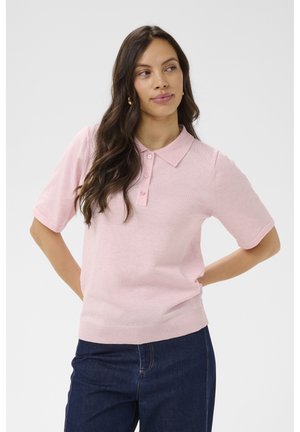 Hosszú, sötét hajú nő világos rózsaszín rövid ujjú polóingben és sötétkék farmerben, kezét a háta mögött tartva áll.