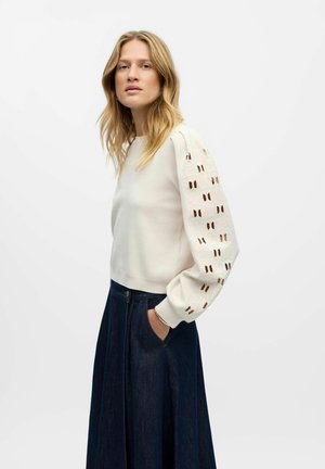 Sweatshirt couleur crème avec des manches texturées et bouffantes présentant des découpes géométriques. Associé à une jupe en denim foncé et des poches avant.