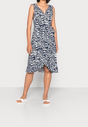 Femme portant une robe portefeuille bleu marine sans manches avec des motifs abstraits blancs et des sandales blanches à enfiler, debout devant un fond uni.