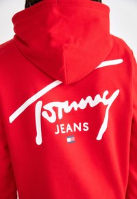 Vista posteriore di una persona che indossa un hoodie rosso di Tommy Jeans con il logo bianco in firma e una piccola bandiera rettangolare sul retro.