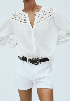 Bílá halenka s dlouhým rukávem s průhlednou krajkovou vsadkou, kombinovaná s bílými krátkými šortkami s ohrnutými lemy a černým páskem s cvočky na stojícím modelu.