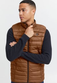 Braune wattierte Steppweste mit Frontreißverschluss und Seitentaschen, kombiniert mit einem marineblauen, gerippten Langarmshirt. Markantes Markenlogo auf der Brust.