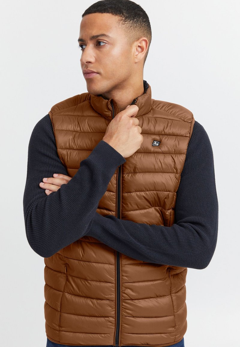 Braune wattierte Steppweste mit Frontreißverschluss und Seitentaschen, kombiniert mit einem marineblauen, gerippten Langarmshirt. Markantes Markenlogo auf der Brust.