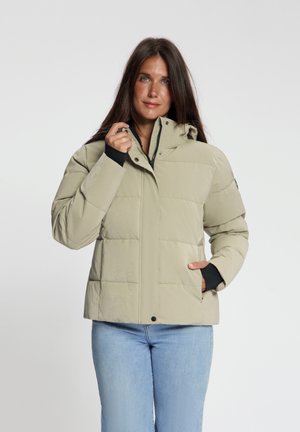 Vrouw draagt een beige donsjacket met capuchon, met één hand de kraag aanpassend, en lichtblauwe spijkerbroek tegen een effen achtergrond.