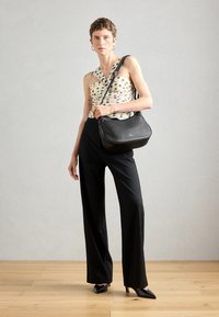 Black leather shoulder bag with a rounded shape and adjustable strap, featuring a subtle logo detail. Model wears a polka dot top and black trousers.