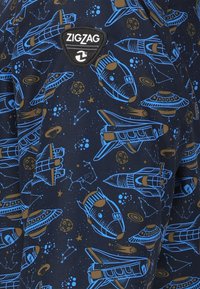 Marinfärgad tyg med ett färgglatt rymdtema som innehåller blå raketer, UFO:n, planeter och stjärnbilder. Zickzack-logotype i svart patch.