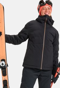 Zwarte geïsoleerde skijack met een hoge kraag, een rits aan de voorkant en zichtbare naden. De persoon houdt oranje skies vast en draagt een roze muts en een skibril.