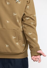Haut à capuche marron avec des motifs d'oiseaux crème, des poignets côtelés et une poche avant. Le matériau semble doux, avec un design global décontracté.