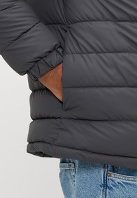 Μαύρο μπουφάν τύπου puffer με ριμπ μανσέτες και οριζόντιες ραφές. Ζεστό, επενδυμένο υλικό. Συνδυασμένο με ανοιχτόχρωμα τζιν παντελόνια.