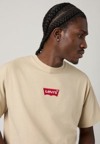 Ung mann med flette i håret iført en beige Levi's T-skjorte, vendt mot høyre mot en ensfarget lys bakgrunn.