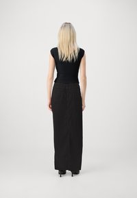 Blond kvinna står med ryggen mot kameran, iklädd en svart topp med kort ärm, lång svart kjol och svarta klackskor framför en enfärgad bakgrund.