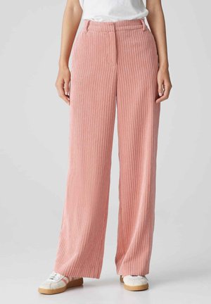 Pantalons en velours côtelé rose clair à jambes larges avec une texture à côtes verticales, taille haute et coupe décontractée, associés à des baskets blanches.
