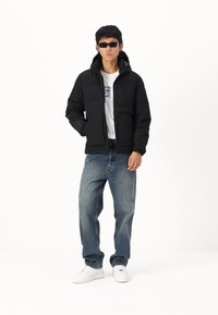 Chaqueta acolchada negra con capucha, cremallera frontal y dos grandes bolsillos delanteros. Combinada con unos vaqueros de mezclilla azul de corte holgado y zapatillas blancas.
