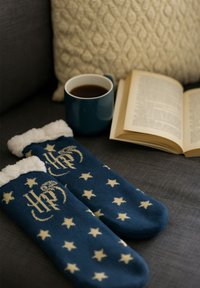Calze di lana blu con stelle color crema e un logo, caratterizzate da un morbido bordo in pelliccia sintetica. In scena ci sono una tazza di caffè e un libro.
