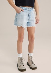 Hellblaue Denim-Shorts mit fransigem Saum, fünf Taschen und Knopfverschluss, getragen mit grauen Schnürstiefeln und weißen Crew-Socken.