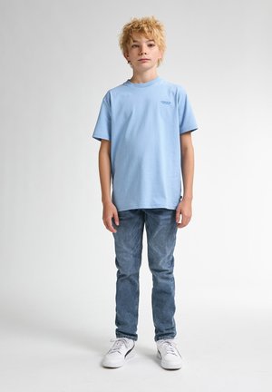 Lichtblauw t-shirt met korte mouwen en een ronde hals, gecombineerd met vervaagde blauwe jeans en witte sneakers, staande op een neutrale achtergrond.