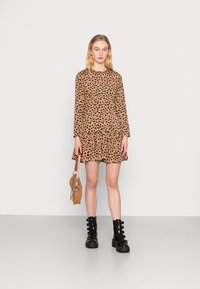 Vestido de manga longa estampado de leopardo com bainha em camadas, combinado com botas pretas com fivela. Modelo segura uma pequena mala castanha. Fundo branco liso.
