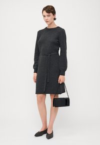 Mørkegrå strikket kjole med lange ermer, tettsittende midje belte og knelengde. Kombinert med en liten svart crossbody-veske og grå leppestifter.