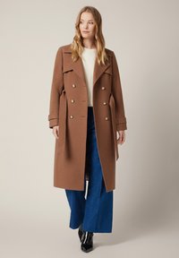 Brązowy wełniany płaszcz dwurzędowy z dużymi guzikami, bocznymi kieszeniami i paskiem w talii, w zestawieniu z jasnym swetrem i niebieskimi spodniami o szerokich nogawkach.