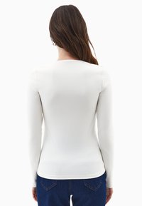 Mujer con cabello largo y marrón, vistiendo una camiseta blanca de manga larga ajustada y jeans azules, mostrada de espaldas contra un fondo blanco.