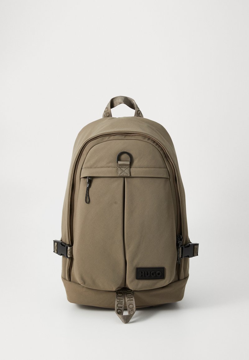 Khaki rugzak met een gladde stofstructuur, een ritsvak aan de voorkant, verstelbare schouderbanden en een zwart logo label. Voorzien van een bovenhandgreep en een gesp.