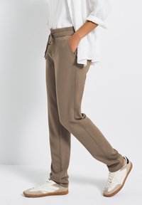 Braune Hose mit elastischem Bund und Seitentaschen, aus strukturiertem Material, kombiniert mit weißen und beigen Sportschuhen. Einfaches, lässiges Design.