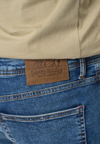Camiseta beige metida dentro de unos vaqueros de denim azul. La etiqueta de cuero en la parte trasera de los vaqueros presenta texto en relieve y acentos decorativos.