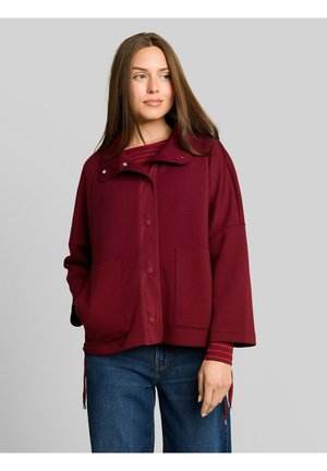 Vrouw die een bordeauxrode jas met knopen draagt over een rood gestreept overhemd en een blauwe spijkerbroek, staand tegen een effen grijze achtergrond.