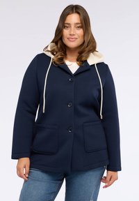 Giacca a vento blu navy con fodera crema, quattro bottoni e due tasche frontali; realizzata in tessuto testurizzato con cordini regolabili.