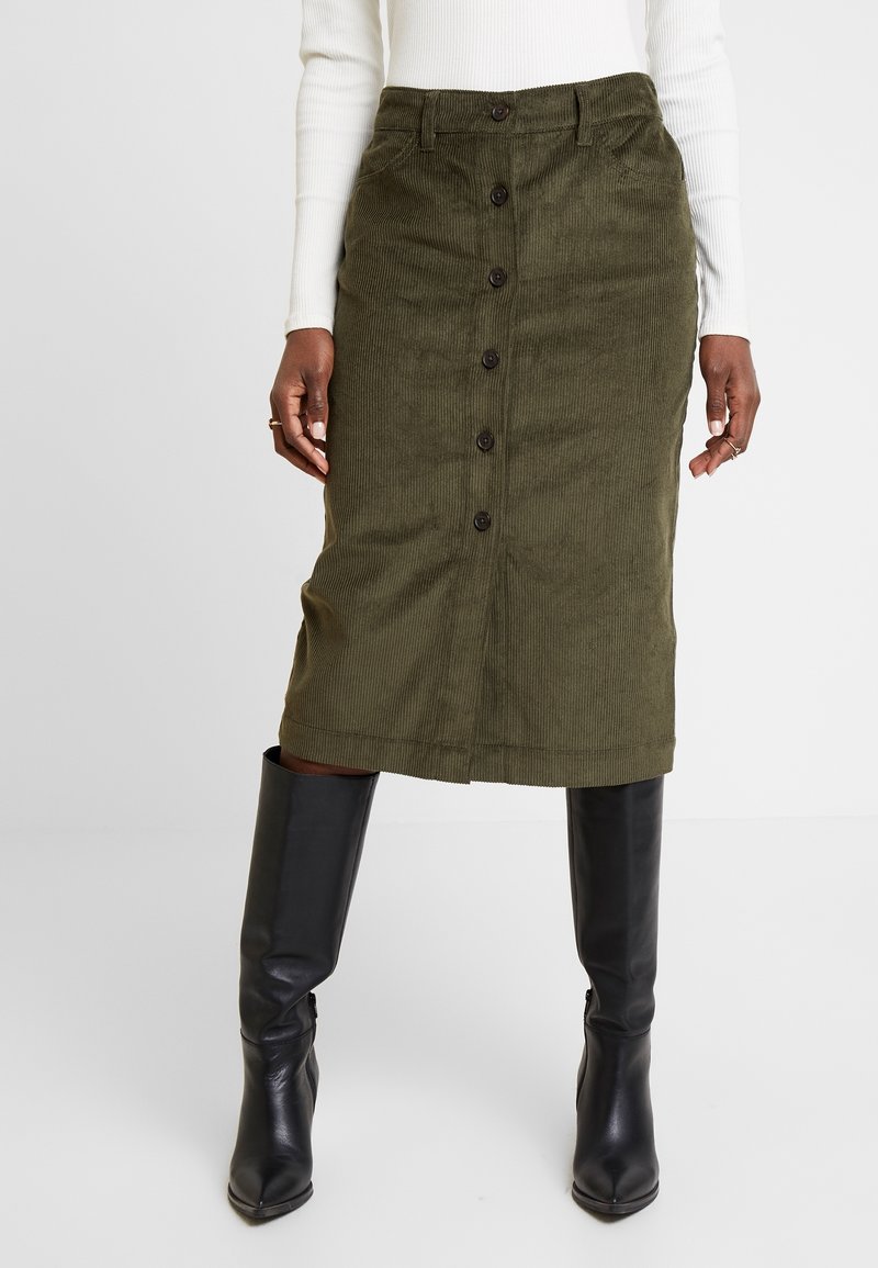Femme portant une jupe midi en velours côtelé vert olive avec boutons sur le devant, des bottes noires montantes et un haut blanc côtelé à manches longues.