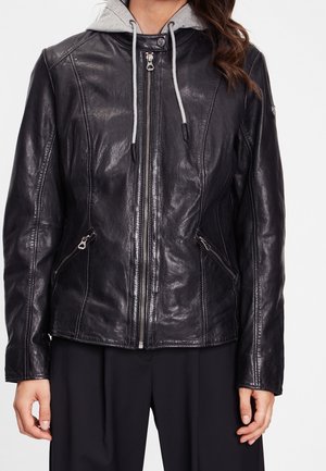 Frau trägt eine schwarze Lederjacke mit Reißverschluss über einem grauen Kapuzenpullover mit Kordeln, kombiniert mit schwarzen Hosen, stehend vor einem schlichten Hintergrund.