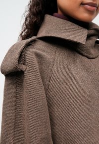 Primer plano de una persona que lleva un abrigo marrón texturizado con un amplio collar y un detalle atado en el hombro sobre un suéter de cuello alto morado oscuro.
