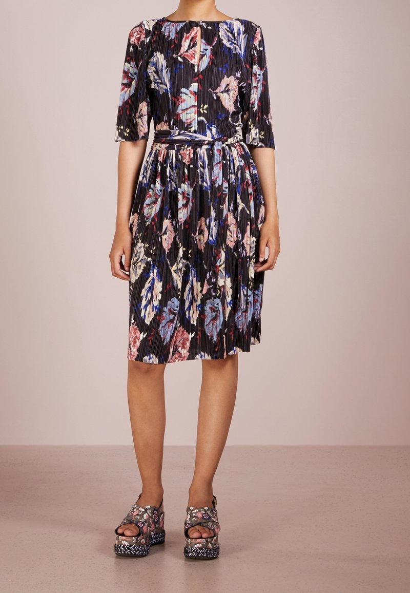 Femme portant une robe noire plissée longueur genou avec un imprimé floral multicolore et des sandales compensées assorties avec des brides à la cheville.