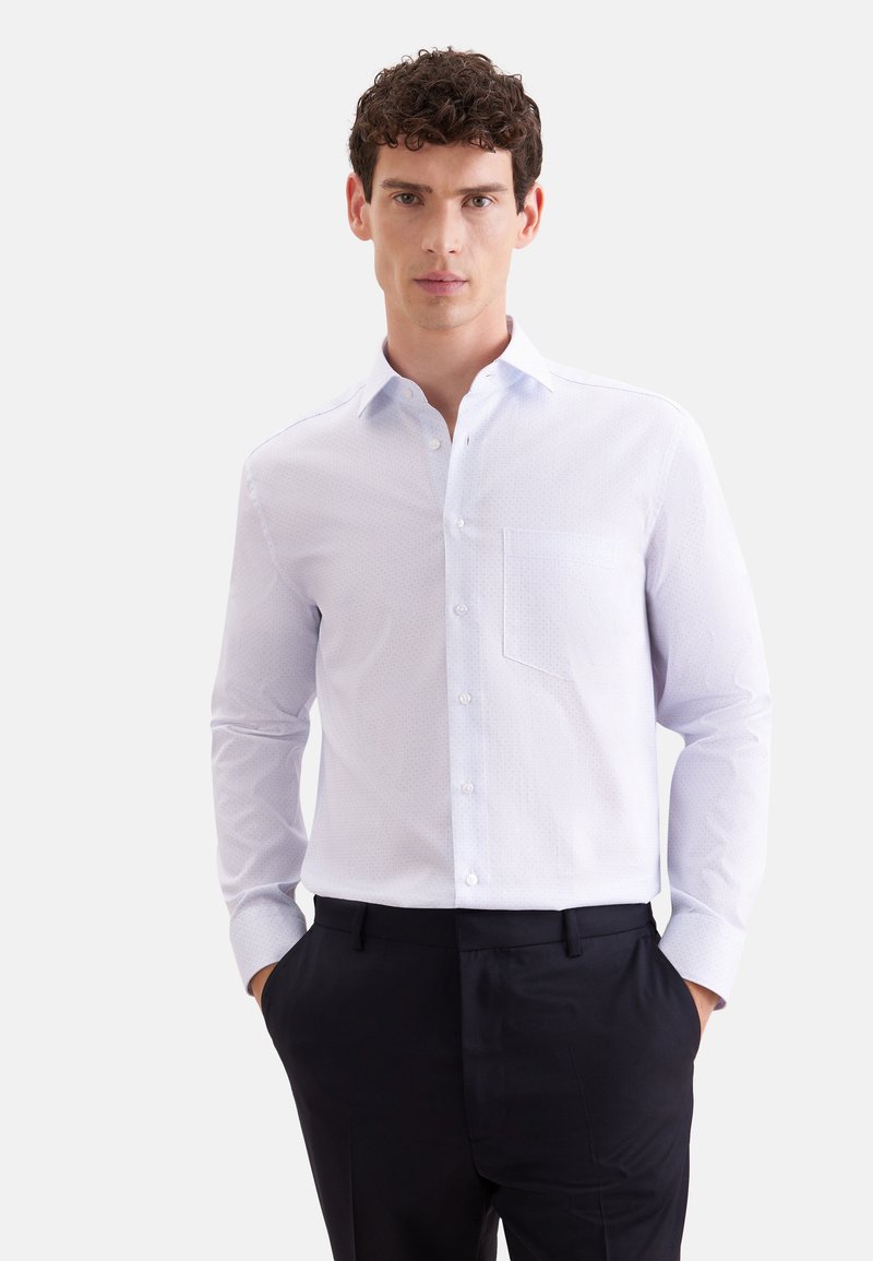 Homme portant une chemise blanche à manches longues avec une poche poitrine et un pantalon noir, debout les mains dans les poches sur un fond blanc.