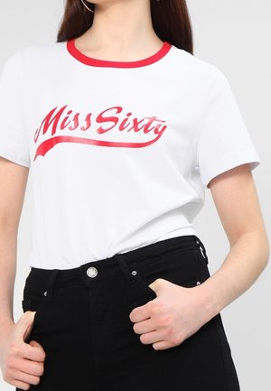 Kvinde iført en hvid t-shirt med rødt kantbånd og "Miss Sixty" tekst, kombineret med sorte bukser med høj talje, hænderne nær lommerne.