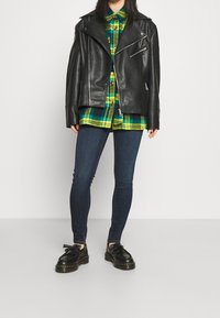 Chaqueta de biker de cuero negro sobre una camisa de cuadros verde y amarillo, combinada con jeans oscuros ajustados y zapatos negros con cordones.