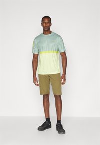 Maglietta sportiva verde chiaro con una sfumatura che sfuma verso un giallo pallido, abbinata a pantaloni corti khaki. Scarpe da ginnastica nere completano l'outfit.