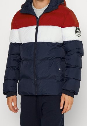 Mężczyzna w pikowanej kurtce z blokami kolorów z czerwonymi, białymi i granatowymi poziomymi paskami oraz naszywką z logo na górnej lewej rękawie.