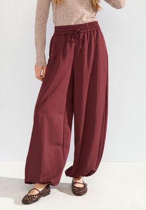 Frau trägt lockere weinrote Hose mit elastischem Bund und Kordelzug, kombiniert mit einem langärmeligen beigefarbenen gestreiften Oberteil und braunen gewebten Ballerinas.