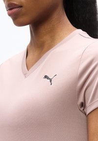 Κοντινό πλάνο ενός ατόμου που φοράει ένα ροζ ανοιχτό μπλουζάκι με V λαιμόκοψη και μαύρο λογότυπο Puma στο στήθος.