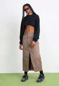 Mujer joven con trenzas largas lleva un sudadera negra cortada, una falda midi beige con abertura frontal, zapatillas negras y gafas de sol sobre un suelo verde.